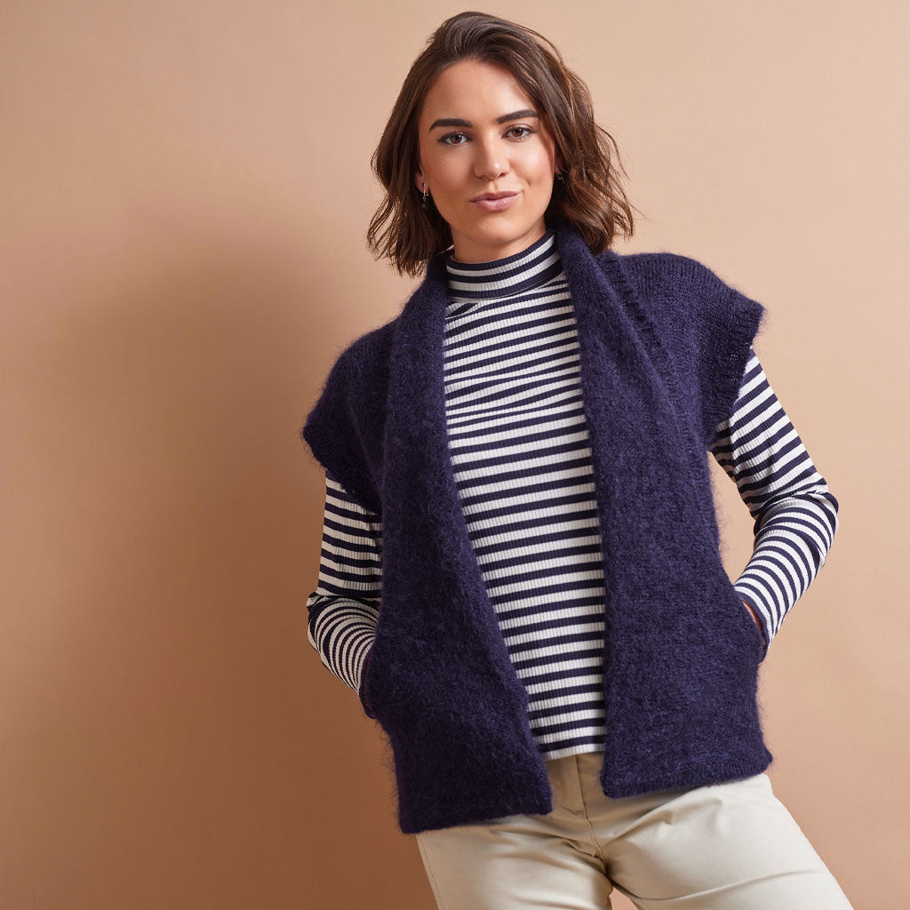 A woman wearing a navy blue fuzzy sleeveless vest over a navy and white striped turtleneck, paired with beige pants, posed against a solid warm beige studio background.