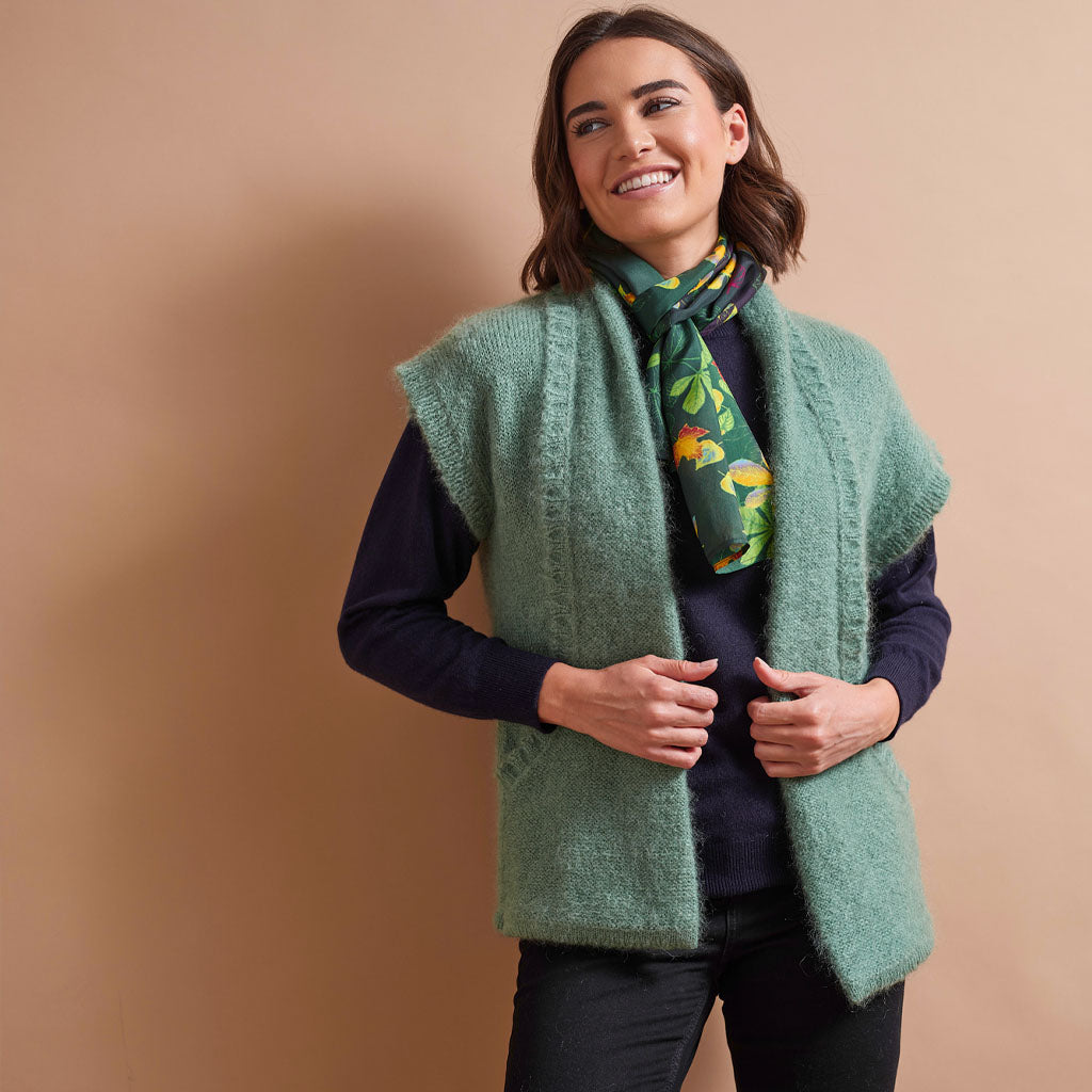A woman wearing a fuzzy sage green sleeveless vest over a dark navy long-sleeve top, accessorized with a green floral scarf, standing against a solid beige background.