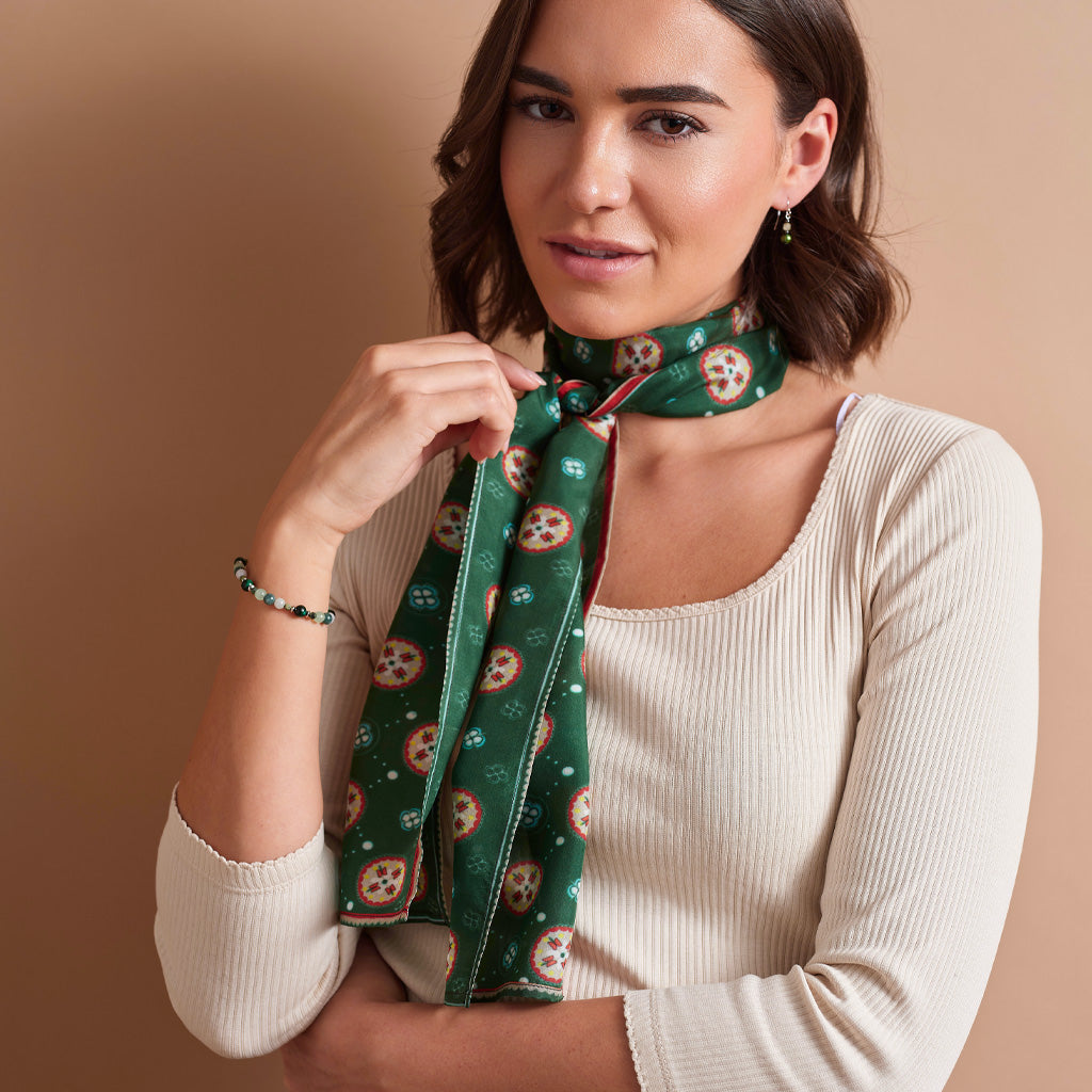 A woman wearing a cream ribbed long-sleeve top and a green patterned silk scarf tied around her neck, posed against a solid warm beige background.