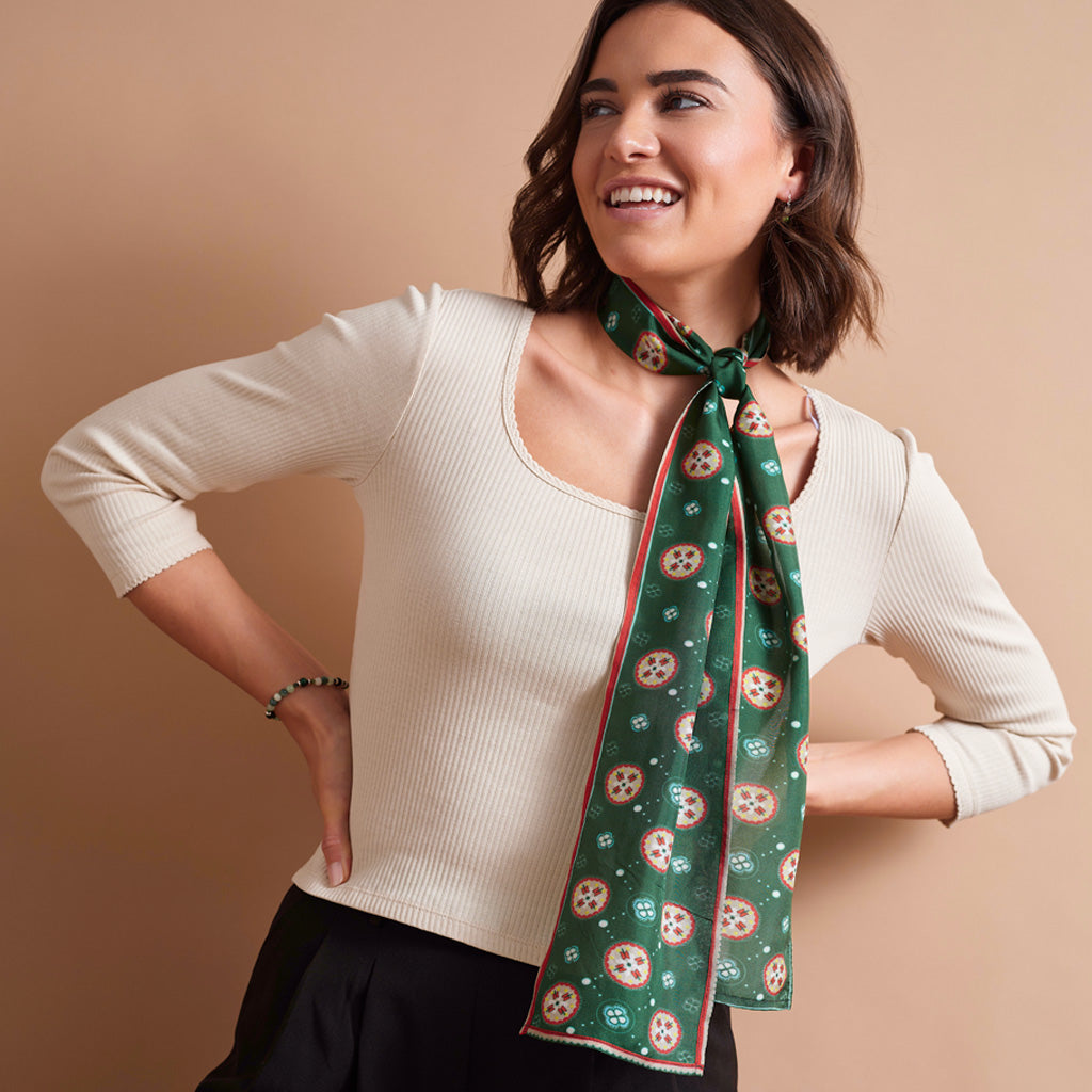 A woman wearing a cream ribbed knit top and black bottoms, styled with a green patterned silk scarf tied around her neck, posed against a solid beige studio background.