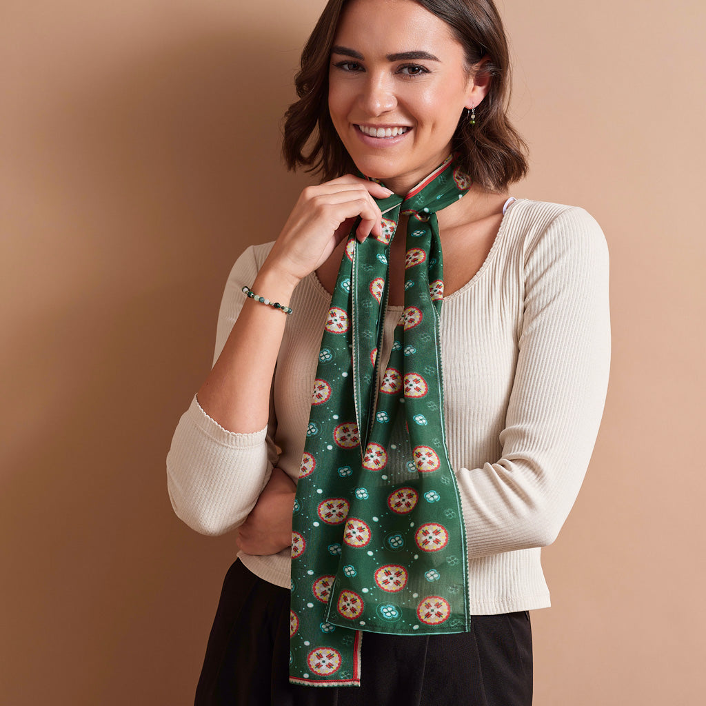 A woman wearing a cream ribbed knit sweater and black skirt models a green silk scarf with a pattern of red and white cherubs and small white stars, tied around her neck against a solid tan background.