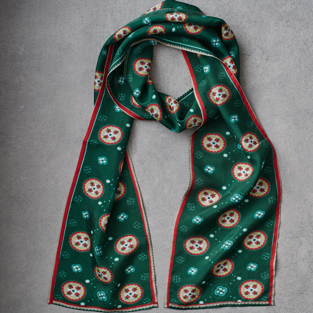 A green silk scarf with a repeating circular medallion pattern in red, white, and gold, finished with a red border, laid flat on a gray textured background.