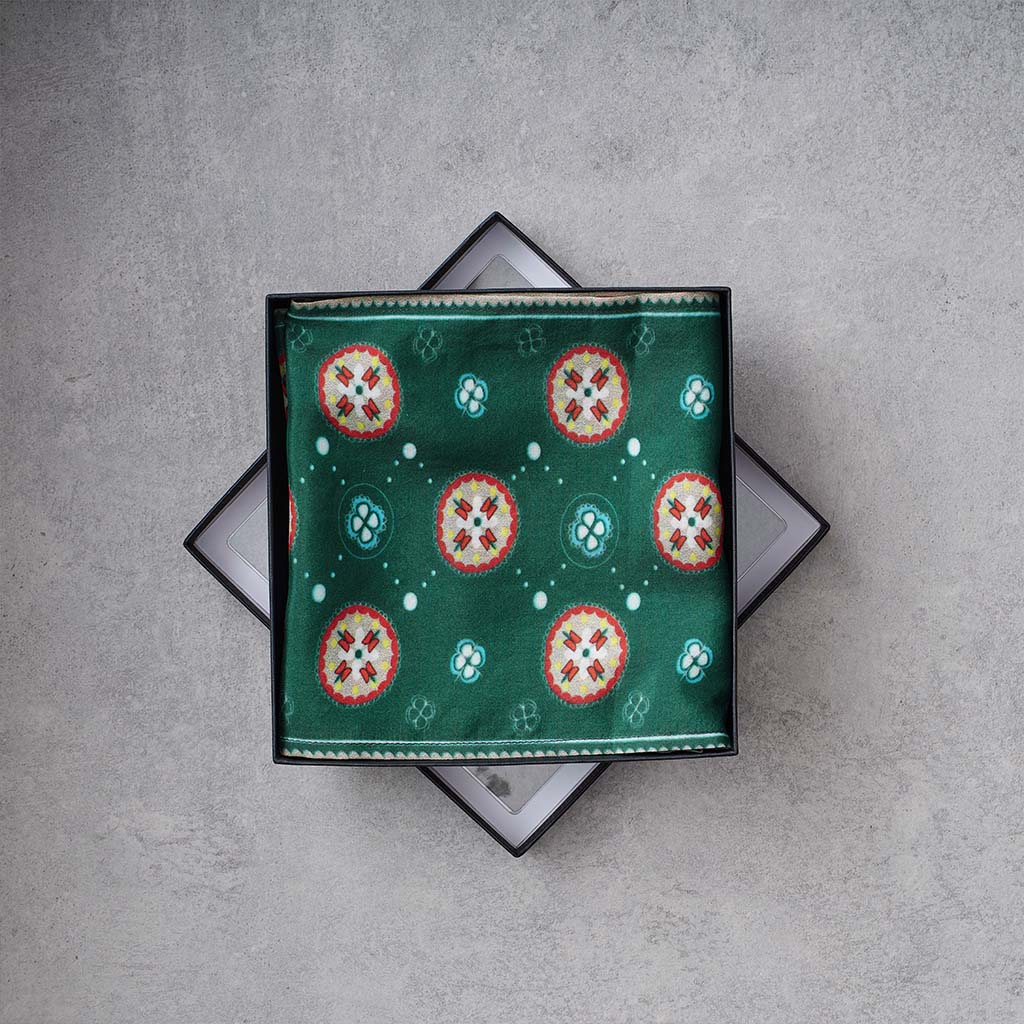 A square green silk-like scarf with a repeating circular medallion pattern in red, white, and teal, presented in an open black gift box on a textured gray concrete surface.