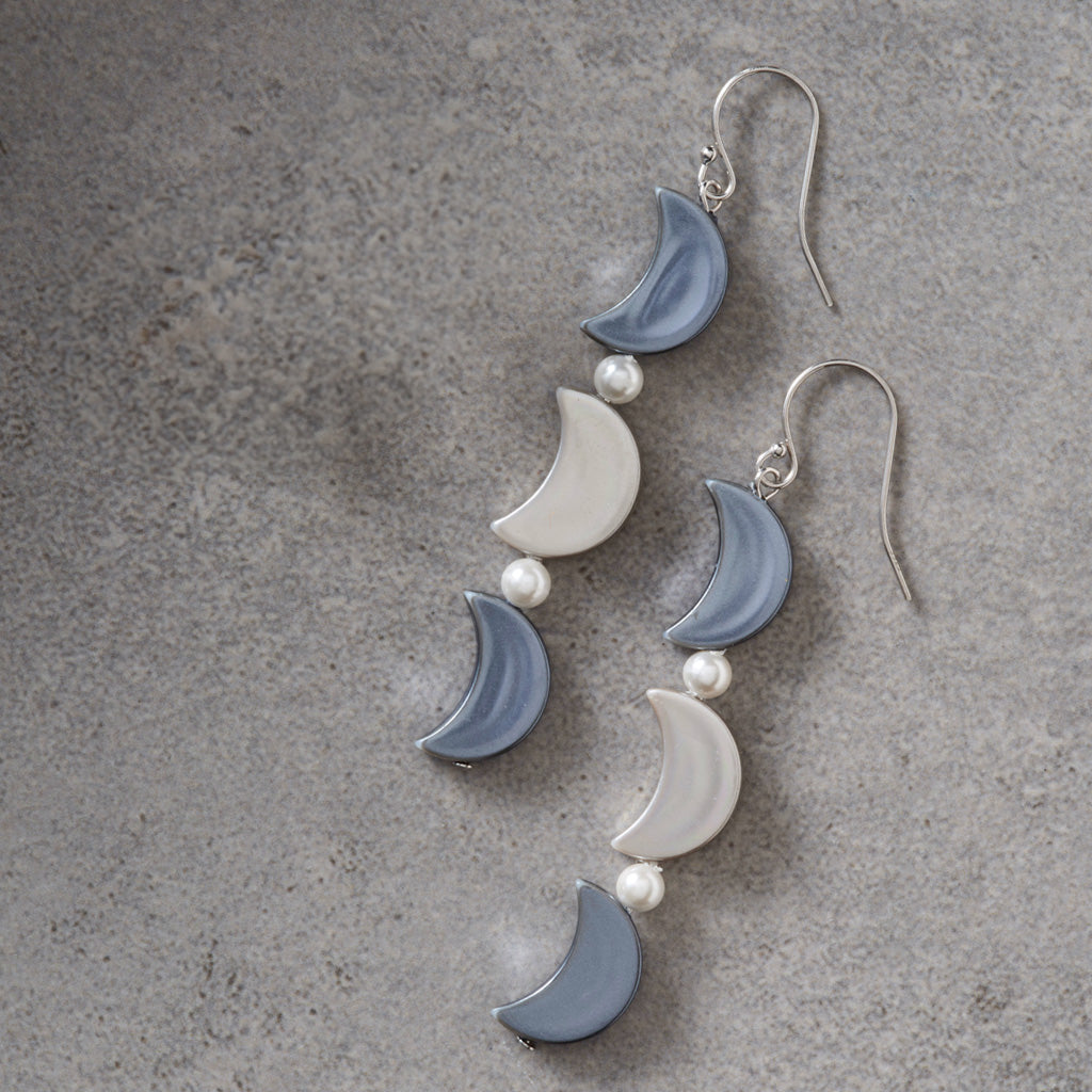 A pair of dangling crescent moon earrings with alternating light blue and white moon charms, separated by small white pearl beads, hanging from silver fishhook wires on a textured gray background.
