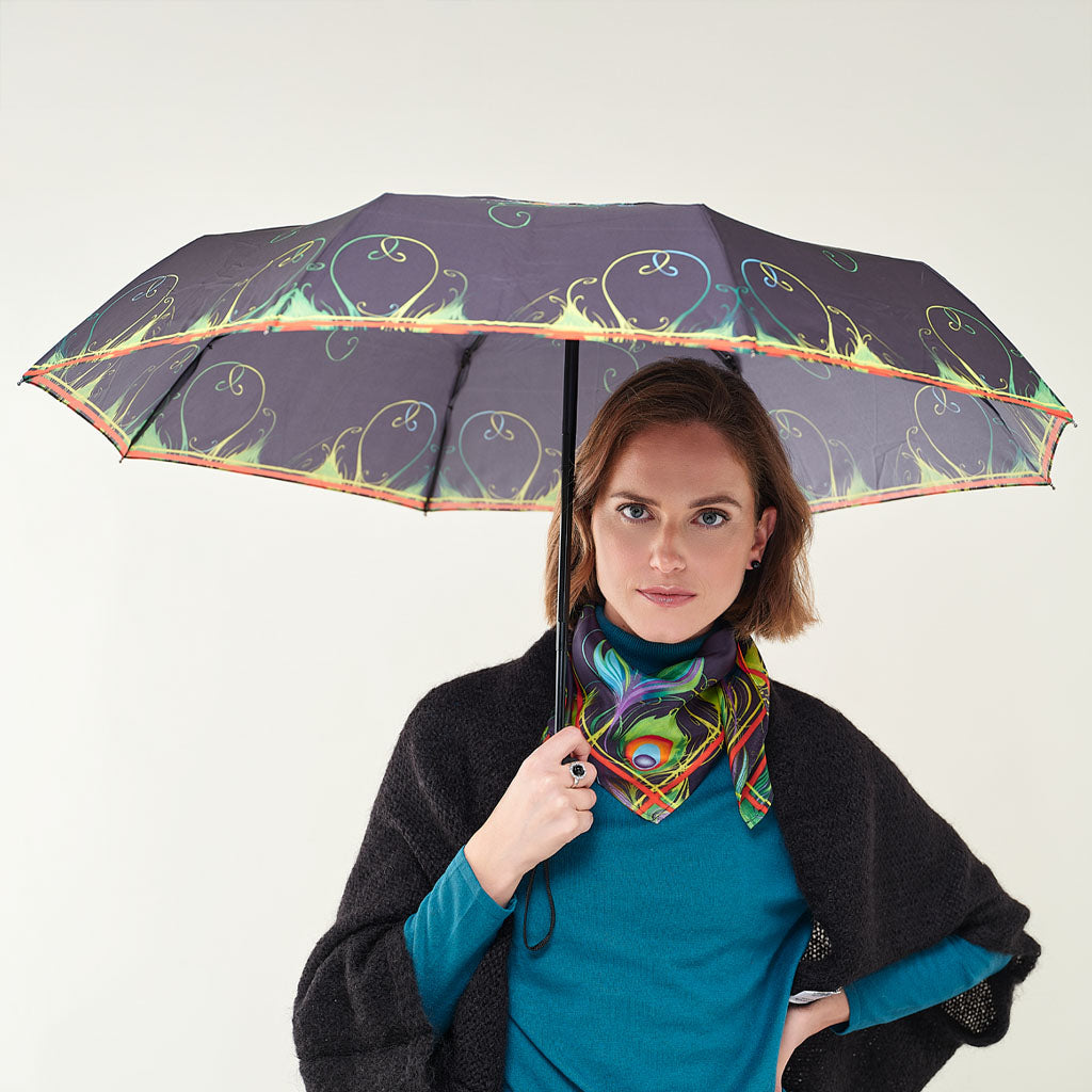 A woman holding a dark purple umbrella with a colorful peacock and floral pattern, wearing a teal top, a black shawl, and a vibrant multicolor scarf against a plain light gray background.