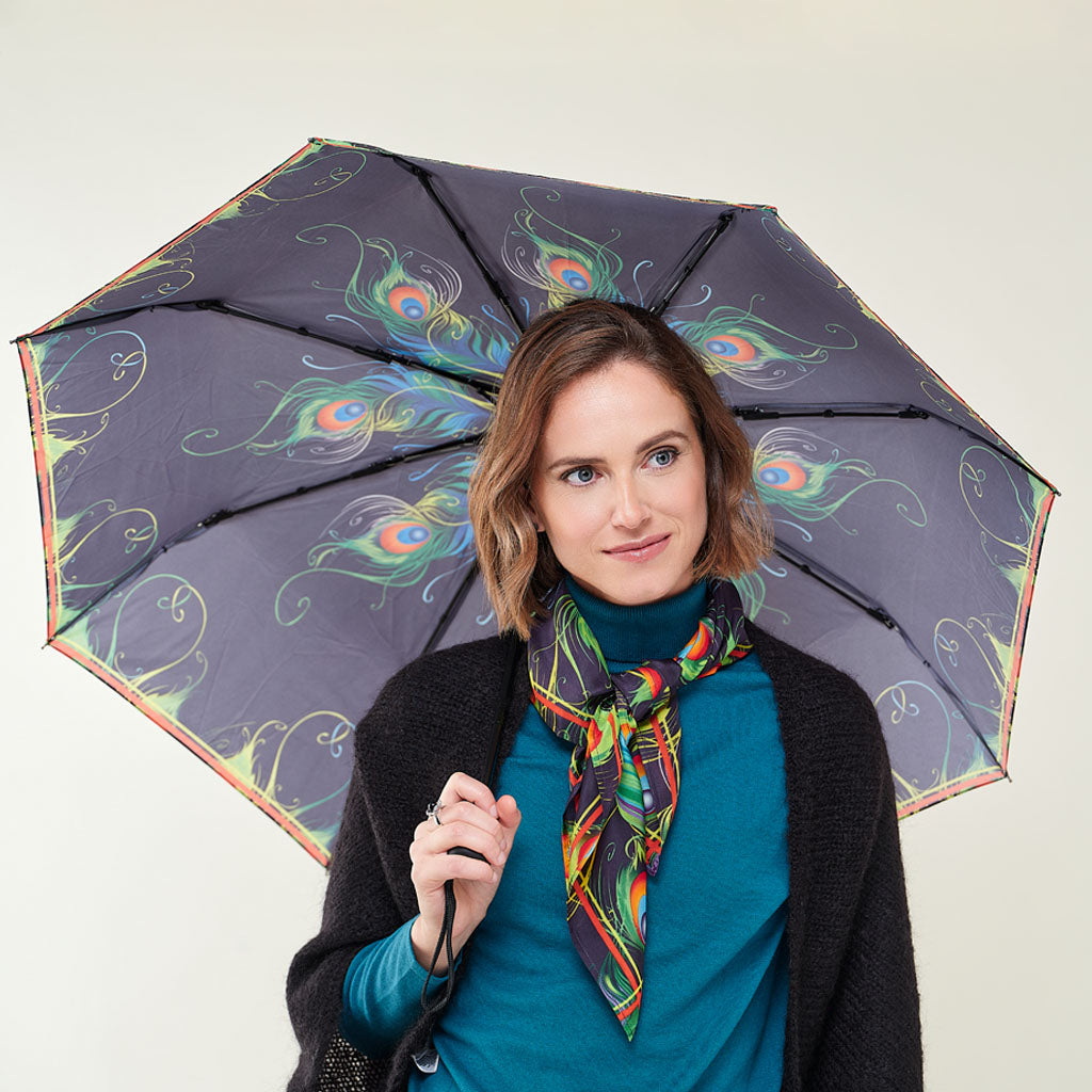 A woman holding a dark gray umbrella with a colorful peacock feather and swirl pattern, wearing a teal turtleneck, a black cardigan, and a matching multicolor patterned scarf against a plain light background.