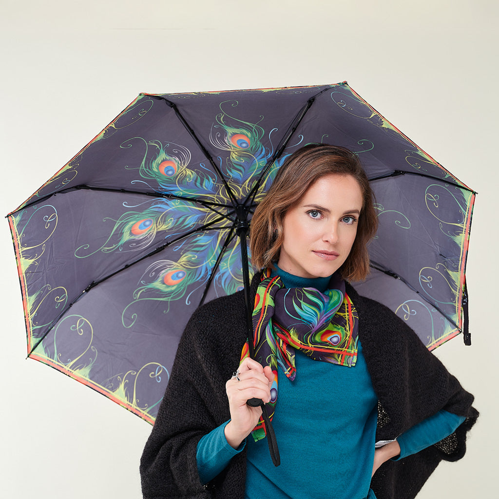A woman holding a dark purple umbrella with a colorful peacock and floral pattern, wearing a teal top, a black shawl, and a vibrant multicolor scarf against a plain light gray background.