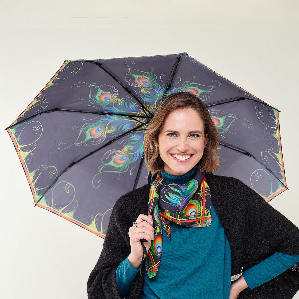 A smiling woman holds up a large open umbrella and wears a matching scarf. The umbrella and scarf feature a dark gray base with a vibrant peacock feather pattern in blue, green, orange, and yellow, set against a plain light beige background.