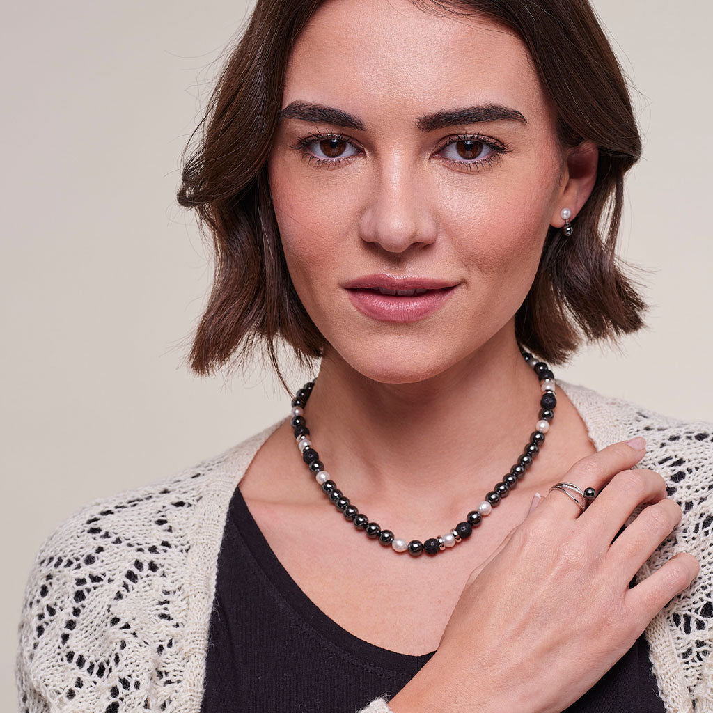 A woman with short brown hair models a beaded necklace with black, white, and pinkish-red beads, paired with a black top and a cream-colored crochet cardigan.