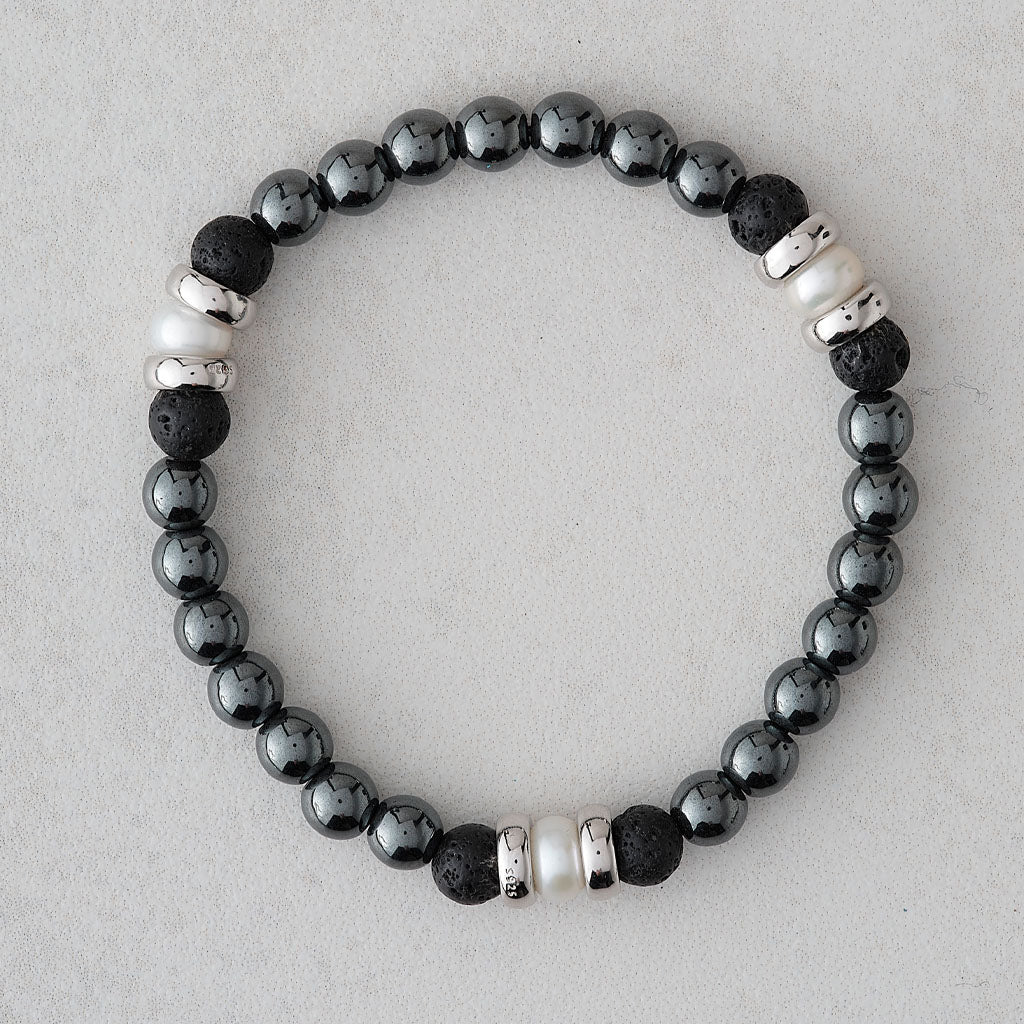 A beaded stretch bracelet featuring black and dark gray volcanic lava beads, white pearl-like beads, and silver-toned spacer beads, displayed on a light gray textured background.