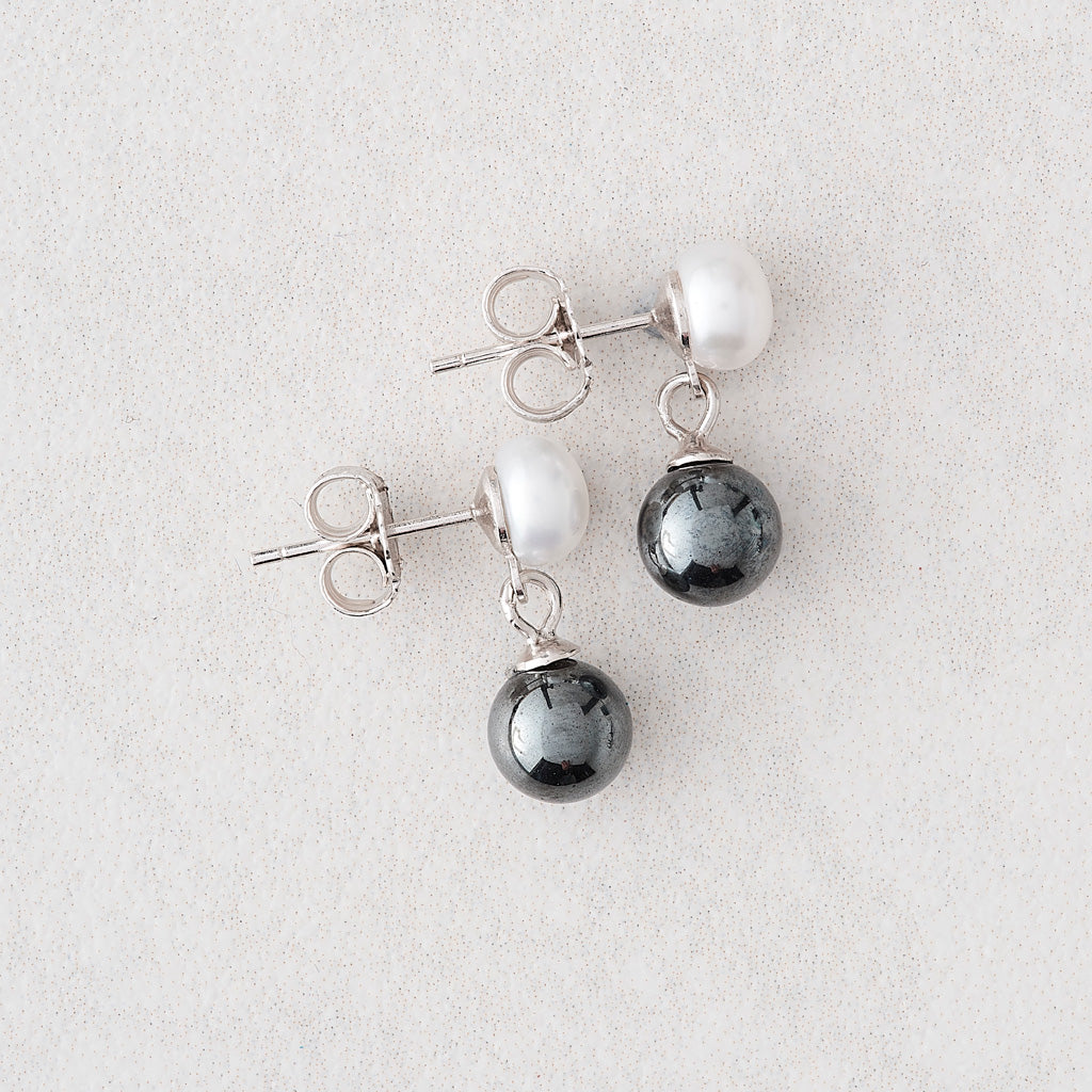 A pair of silver stud earrings featuring a white pearl and a dark gray teardrop-shaped pearl, displayed on a light gray textured background.
