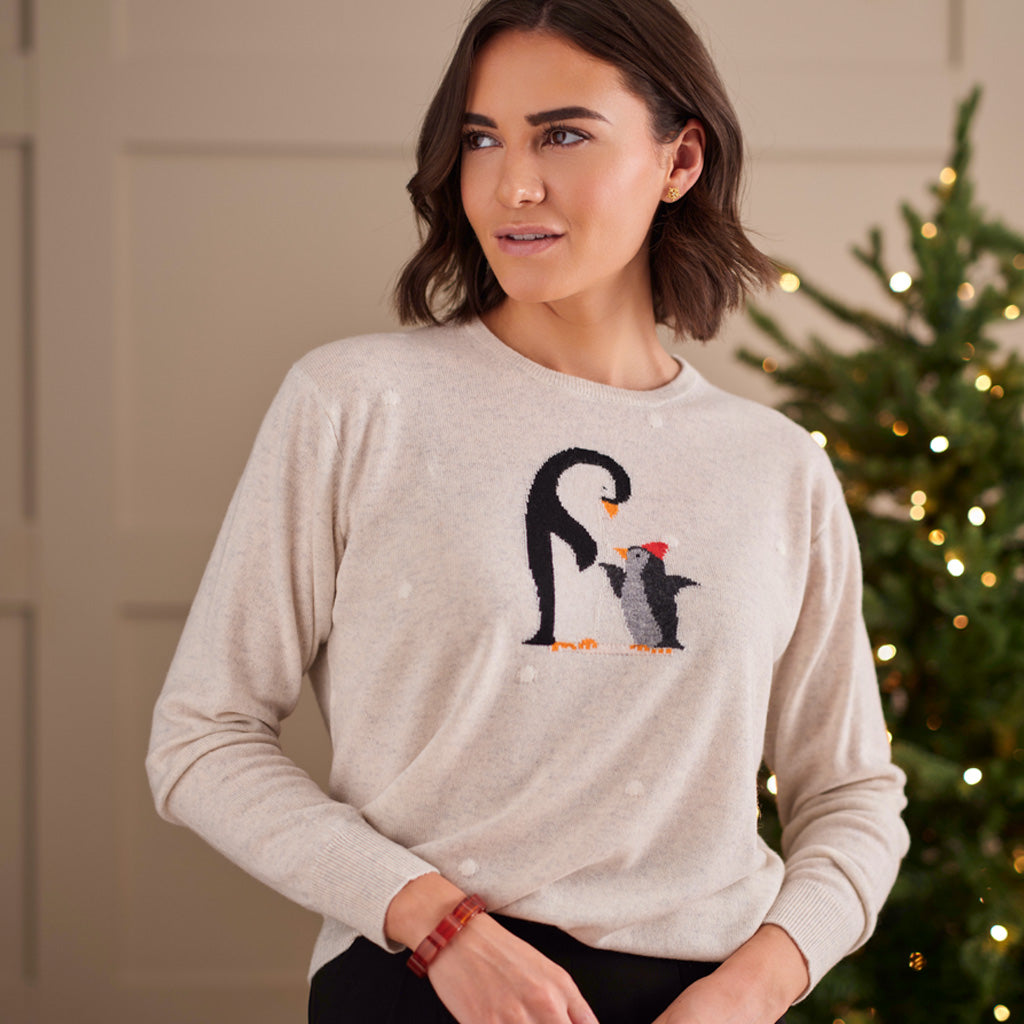A woman wearing a light beige crewneck sweatshirt with a penguin graphic, posed in front of a softly lit Christmas tree.