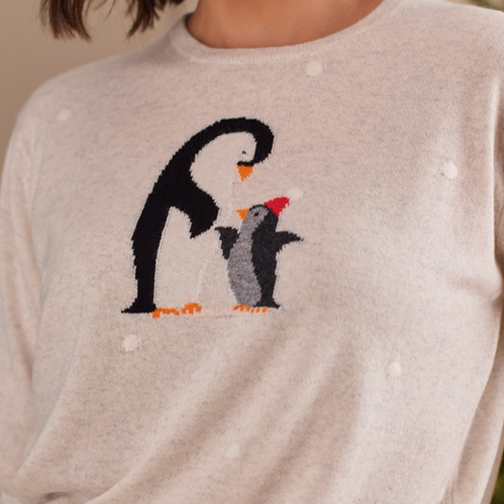 A close-up of a person wearing a light beige knit sweater with a penguin design on the chest. The sweater features a large black and white penguin with orange feet and beak, and a smaller gray penguin with a red beak, accented with small white dot details.