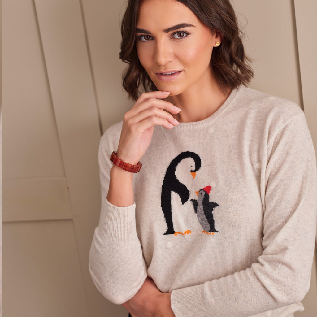 A woman wearing a light beige crewneck sweater with a knitted penguin design on the chest, posed against a neutral beige paneled background.