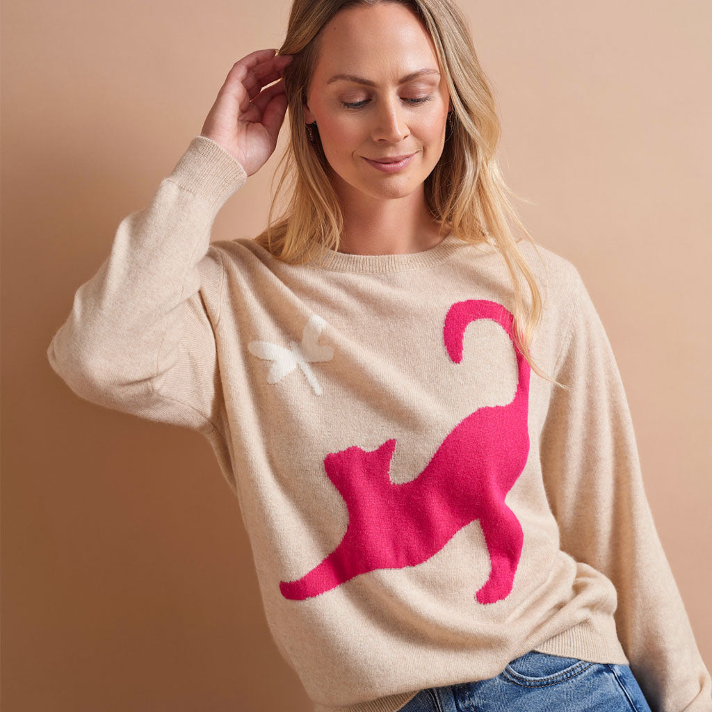 A woman wearing a beige knit sweater with a large hot pink cat silhouette and a small white dragonfly motif, posed against a solid warm beige background.