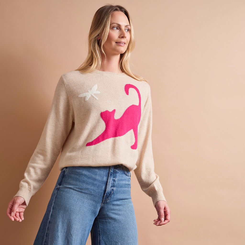 A woman wearing a beige knit sweater with a large hot pink cat graphic and a small white dragonfly motif, paired with blue jeans, against a solid beige studio background.