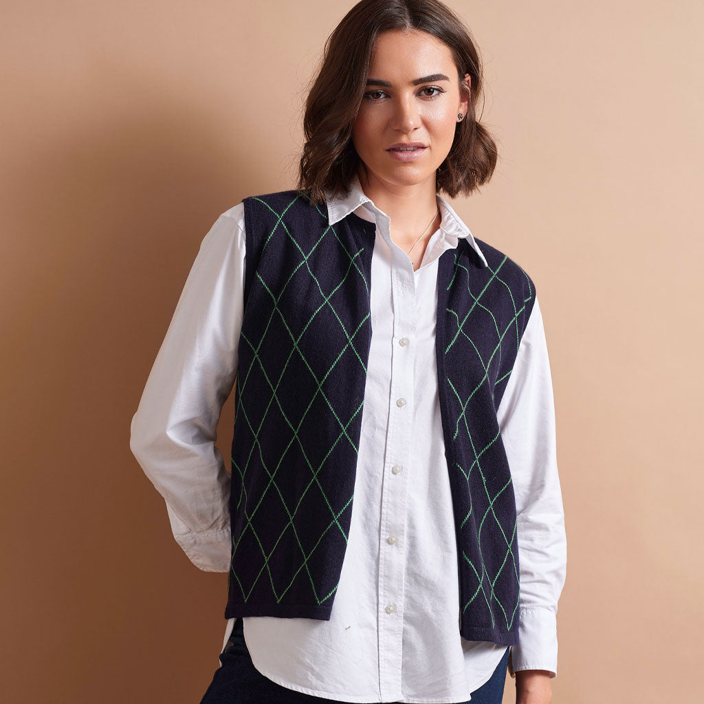 A woman wearing a white button-down shirt and a dark navy vest with a green argyle pattern, posed against a solid beige background.
