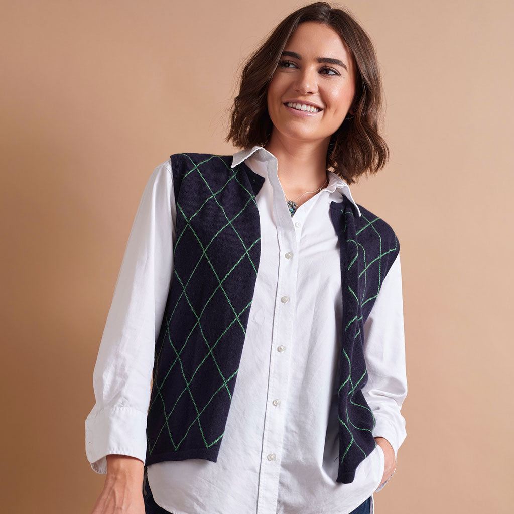 A woman wearing a white button-down shirt and a dark navy vest with a green argyle pattern, posed against a solid beige background.