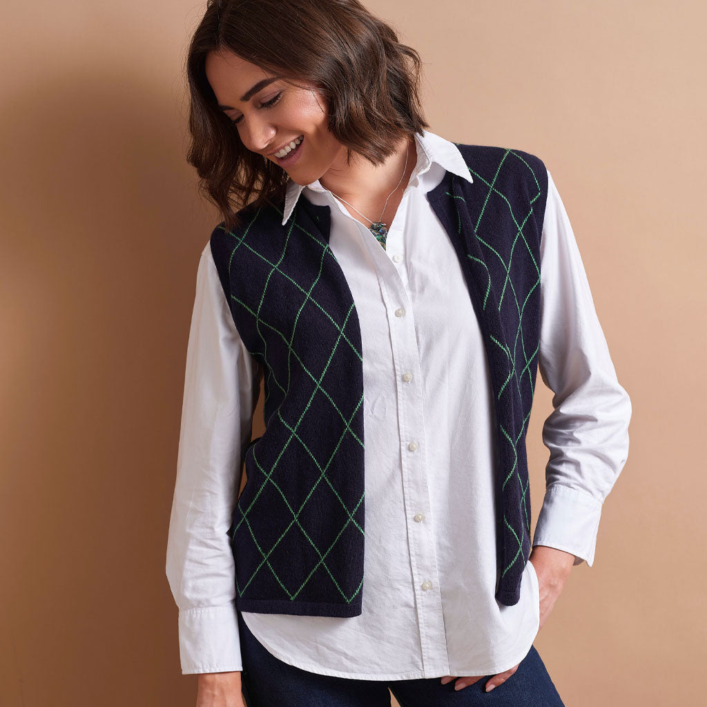 A woman wearing a white button-down shirt and a dark navy argyle vest with green diamond lines, posed against a solid tan background.