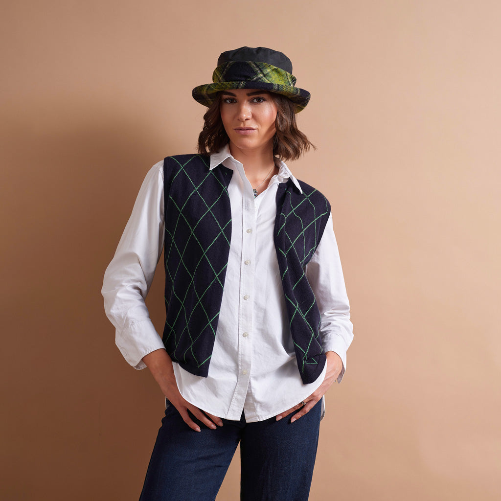 A woman wearing a white button-down shirt and a dark navy vest with a green argyle pattern, posed against a solid beige background.