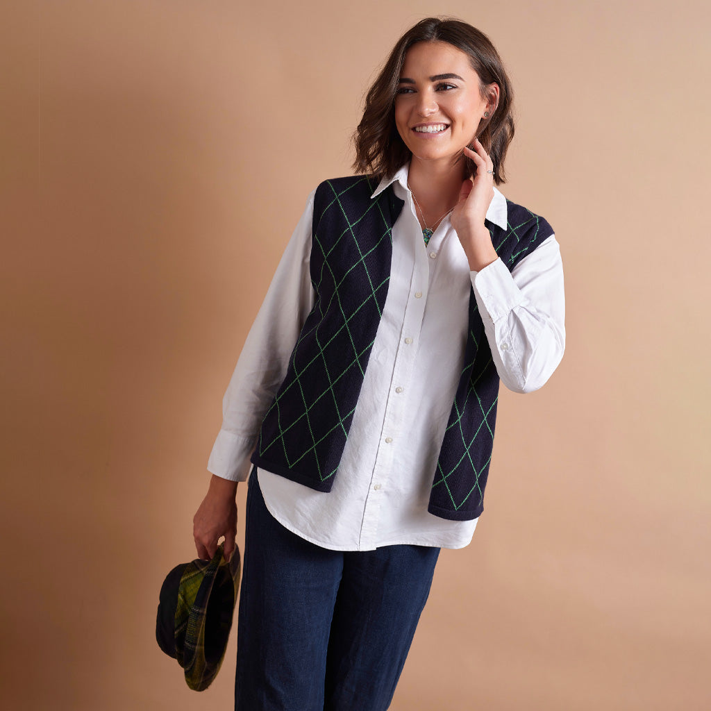A woman wearing a white button-down shirt and a dark navy vest with a green argyle pattern, posed against a solid beige background.