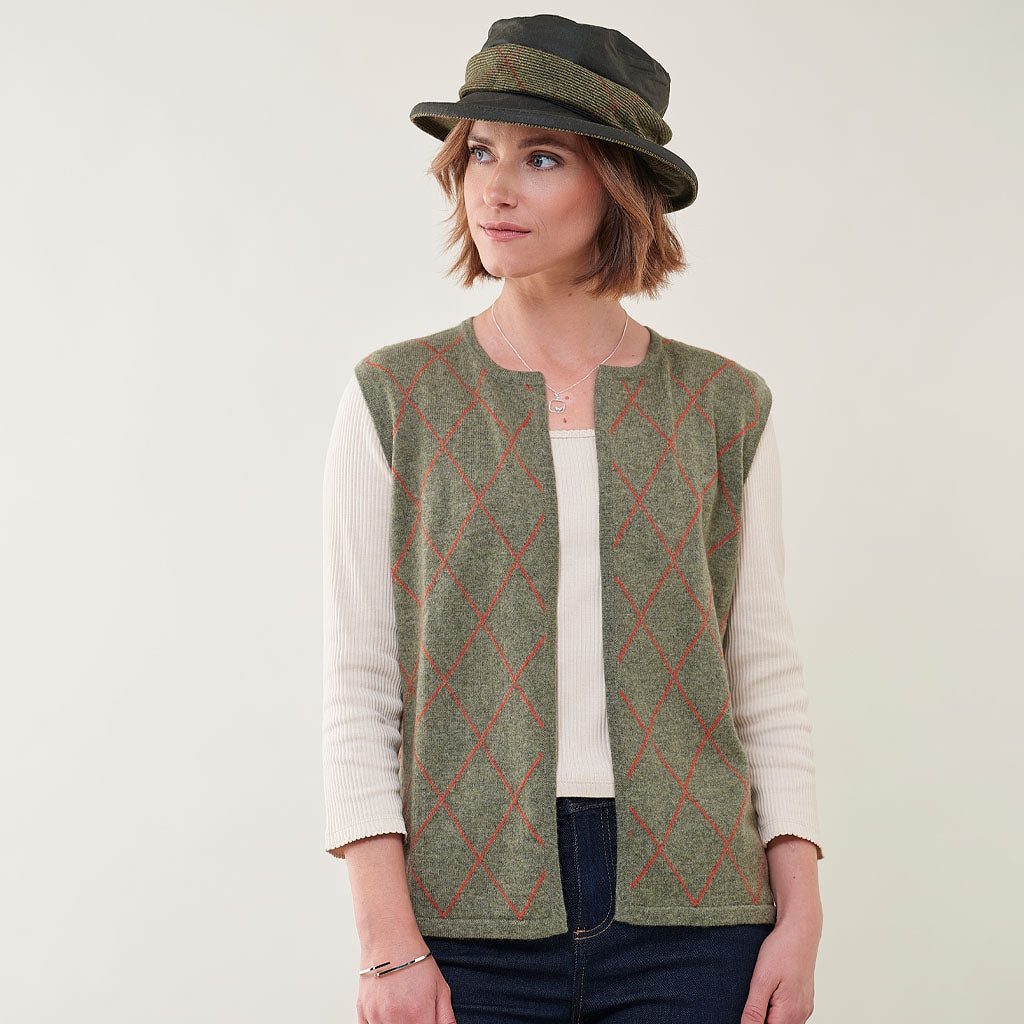 A woman wearing a sleeveless green knit vest with a red argyle pattern over a cream ribbed long-sleeve top, paired with a dark olive green hat, against a plain light background.