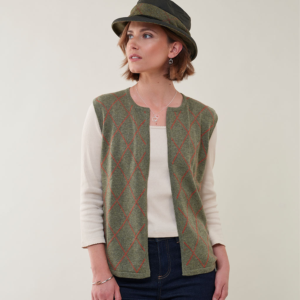 A woman wearing a sleeveless green knit vest with a red argyle pattern over a cream ribbed long-sleeve top, paired with a dark olive green hat, against a plain light background.