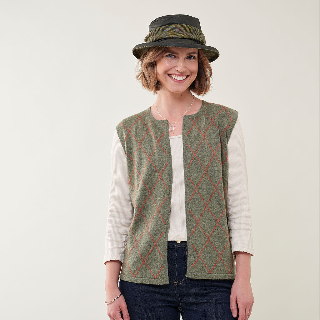 A woman wearing a sleeveless green knit vest with a red argyle pattern over a cream ribbed long-sleeve top, paired with a dark olive green hat, against a plain light background.