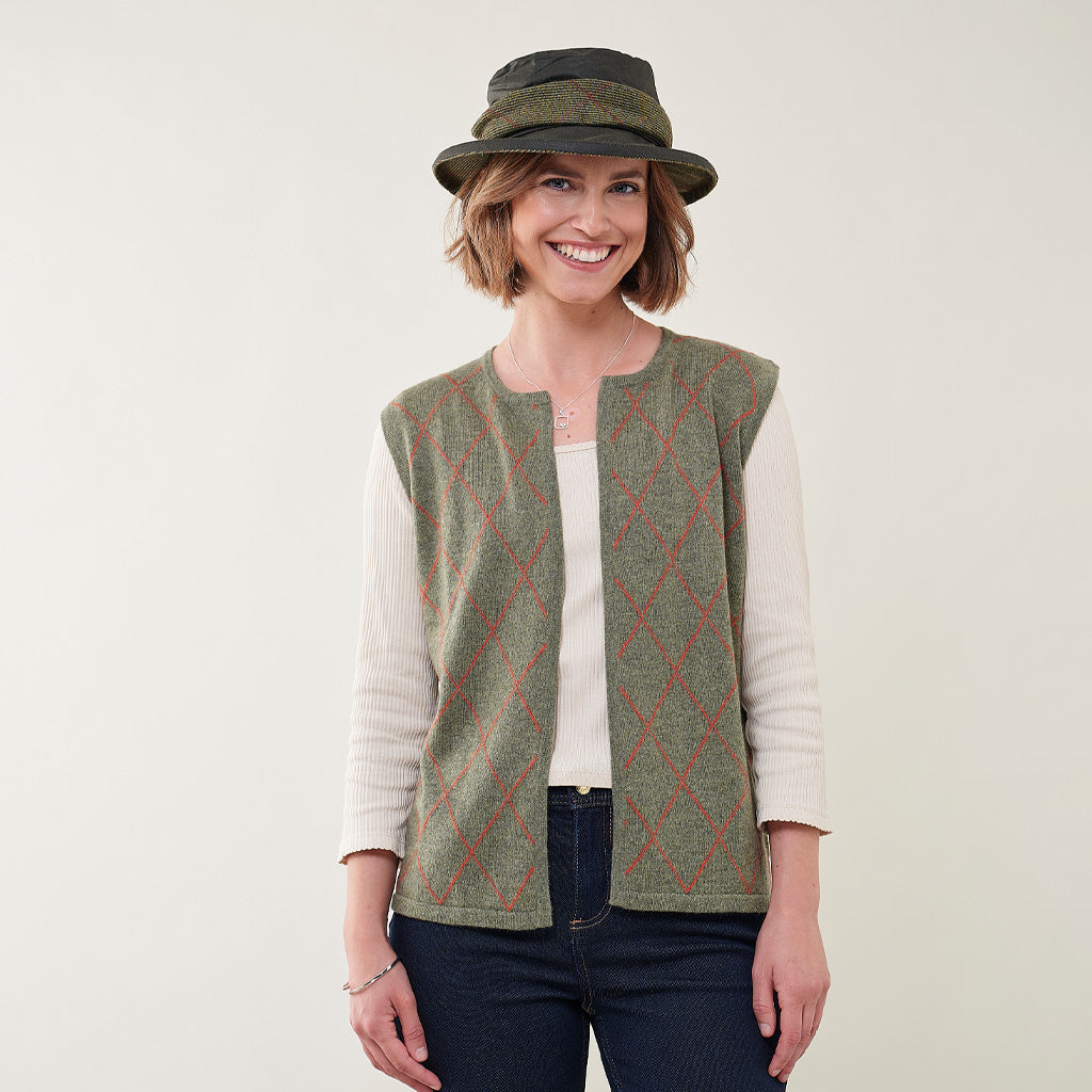 A woman wearing a sleeveless green knit vest with a red argyle pattern over a cream ribbed long-sleeve top, paired with a dark olive green hat, against a plain light background.