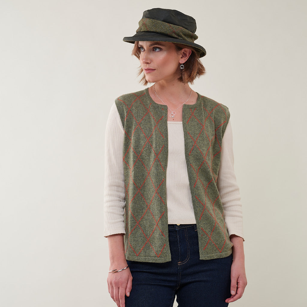 A woman wearing a sleeveless green knit vest with a red argyle pattern over a cream ribbed long-sleeve top, paired with a dark olive green hat, against a plain light background.