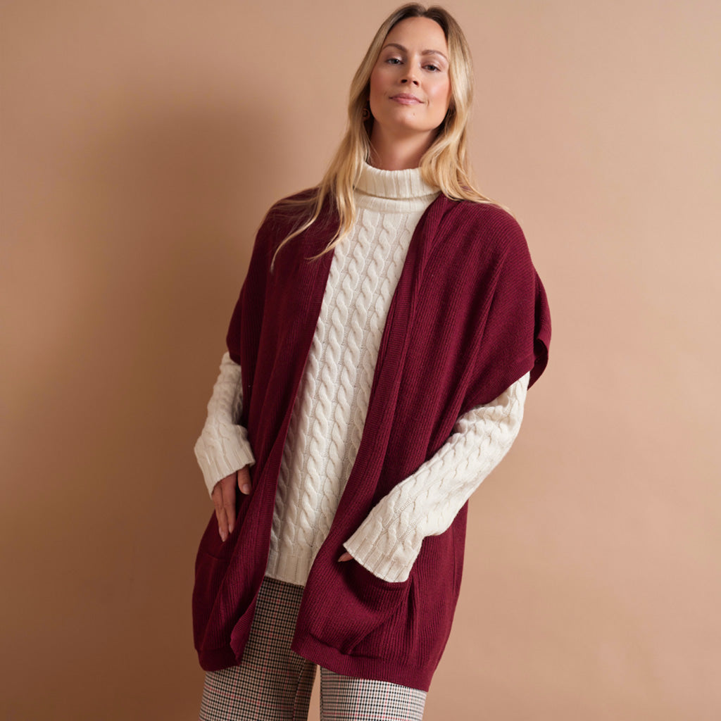 A woman modeling a burgundy open-front knit poncho cardigan over a cream cable-knit turtleneck sweater, paired with gray and white plaid trousers, against a solid warm beige studio background.