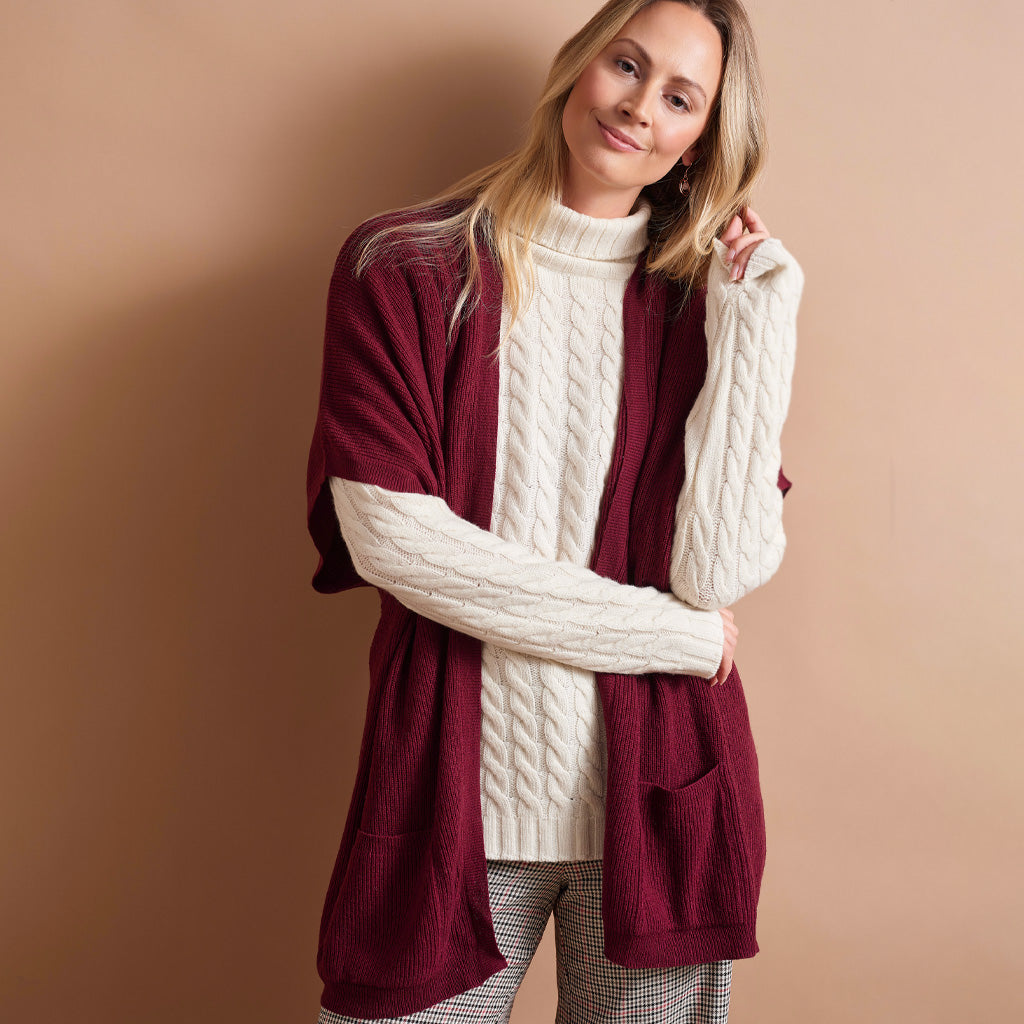 A woman modeling a burgundy open-front cardigan with cream cable-knit sleeves and a cream turtleneck, paired with gray plaid pants, against a solid warm beige background.