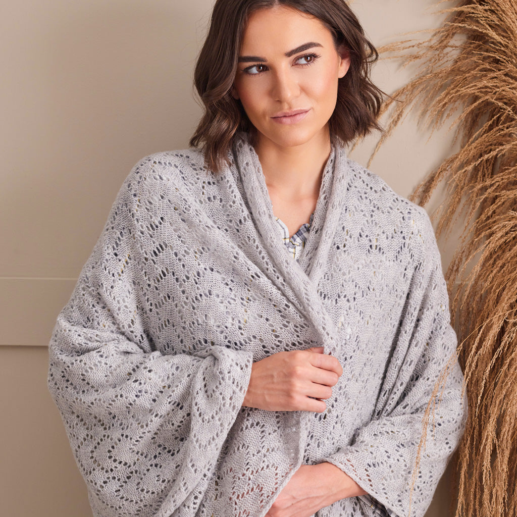 A woman wearing a light gray open-knit shawl wrap, posed next to dried pampas grass against a neutral beige background.