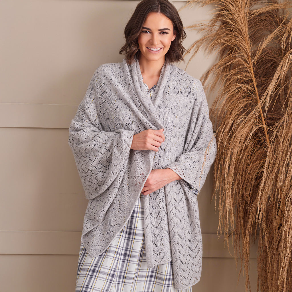 A woman wearing a light gray open-knit shawl wrap draped over her shoulders, paired with a plaid skirt, standing against a beige paneled wall with dried pampas grass on the right.
