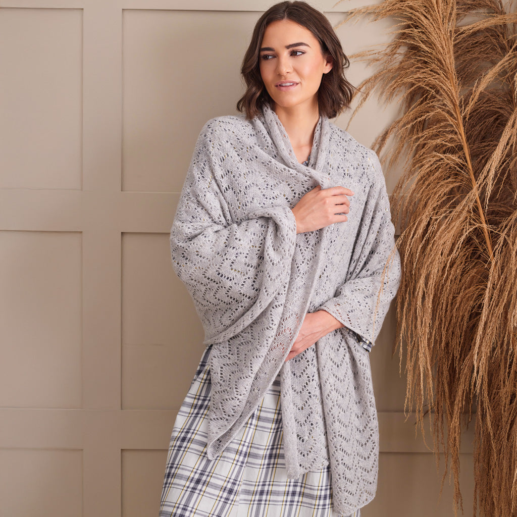 A woman modeling a light gray open-knit shawl draped over her shoulders, paired with a plaid skirt, standing against a beige paneled wall with dried pampas grass on the right.