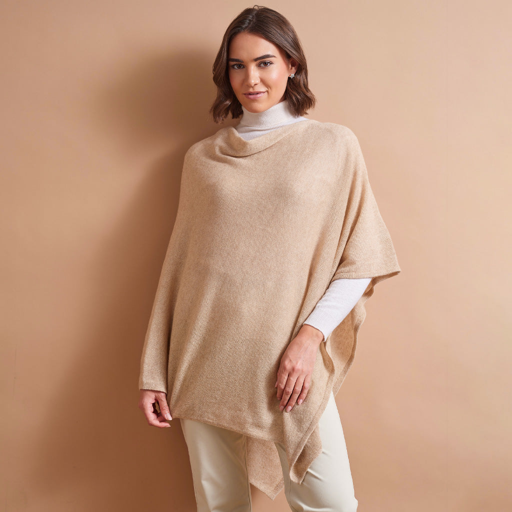 A woman wearing a beige cowl-neck poncho over a white turtleneck and cream pants, standing against a solid beige studio background.