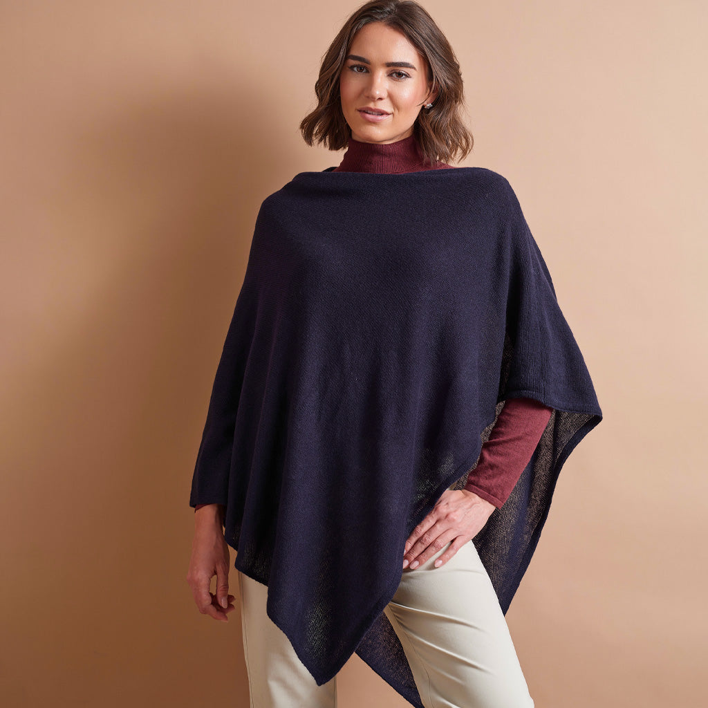 A woman modeling a dark navy blue knit poncho over a burgundy turtleneck and cream pants, set against a solid warm beige studio background.