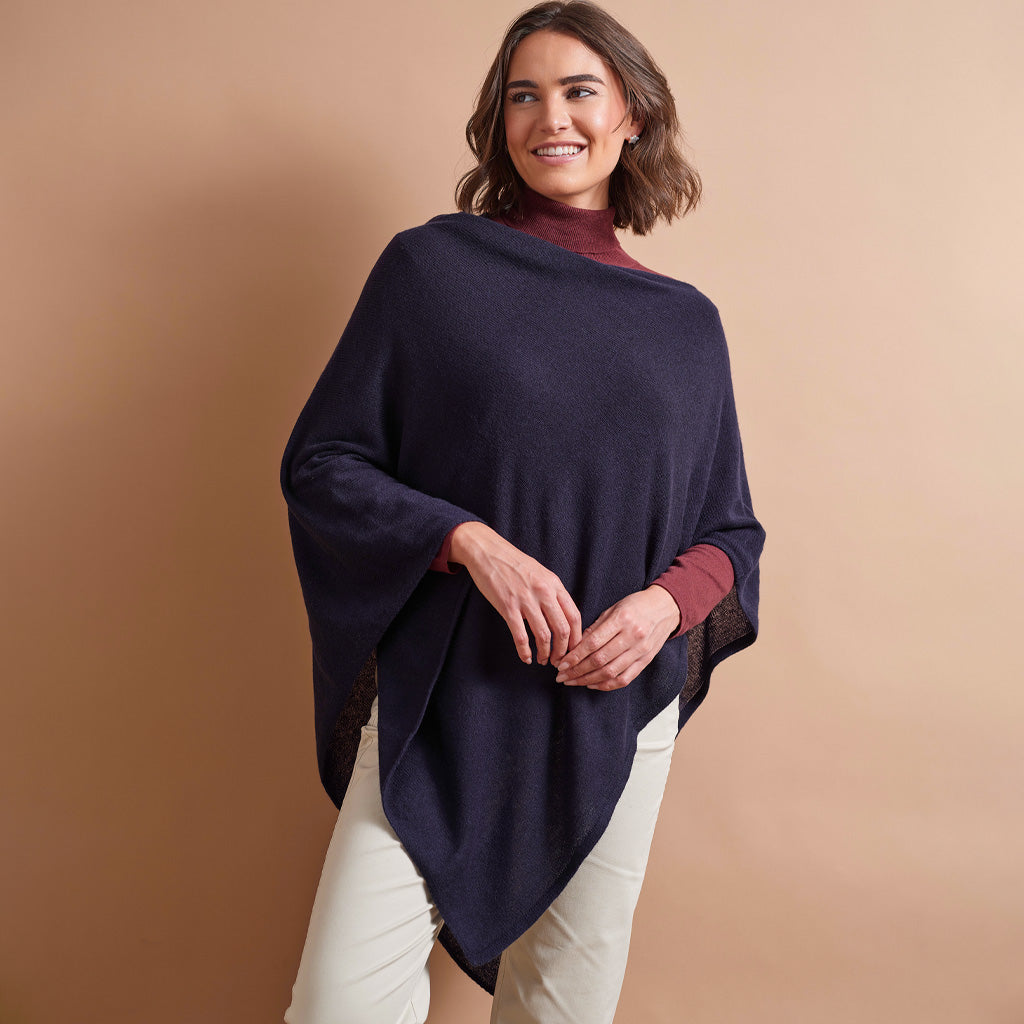 A woman modeling a dark navy blue knit poncho over a burgundy turtleneck and cream pants, set against a solid warm beige studio background.