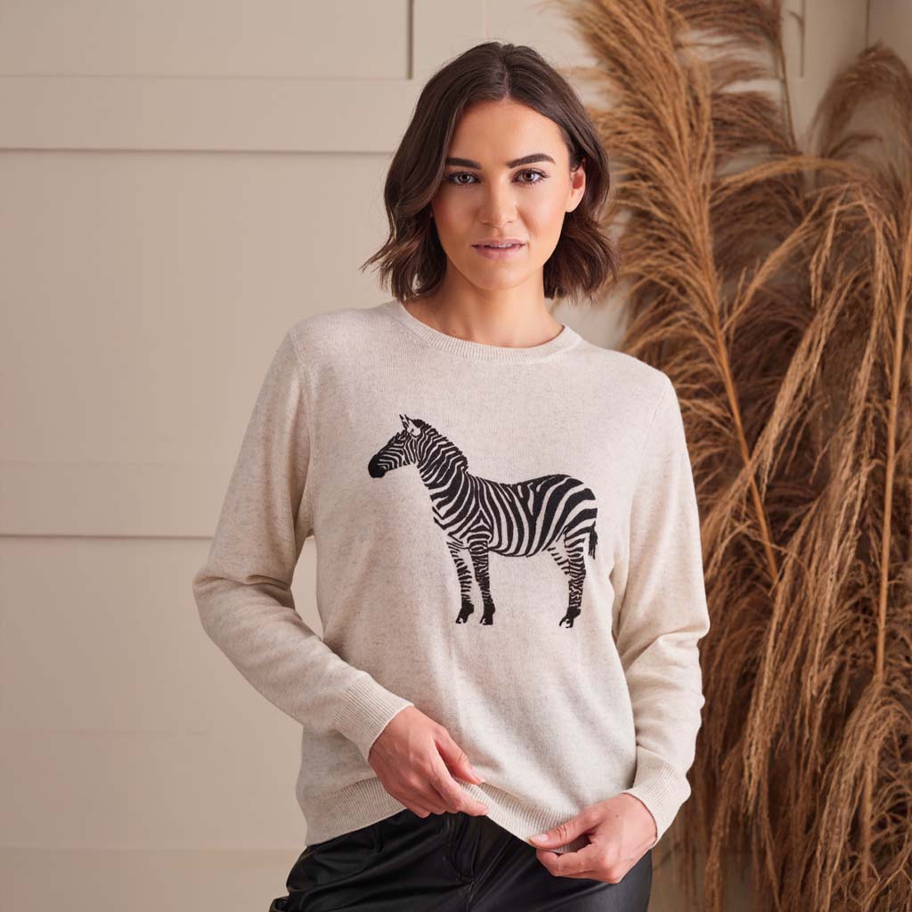 A woman wearing a light beige crewneck sweater with a black and white zebra graphic on the front, standing in front of a neutral wall with dried pampas grass.