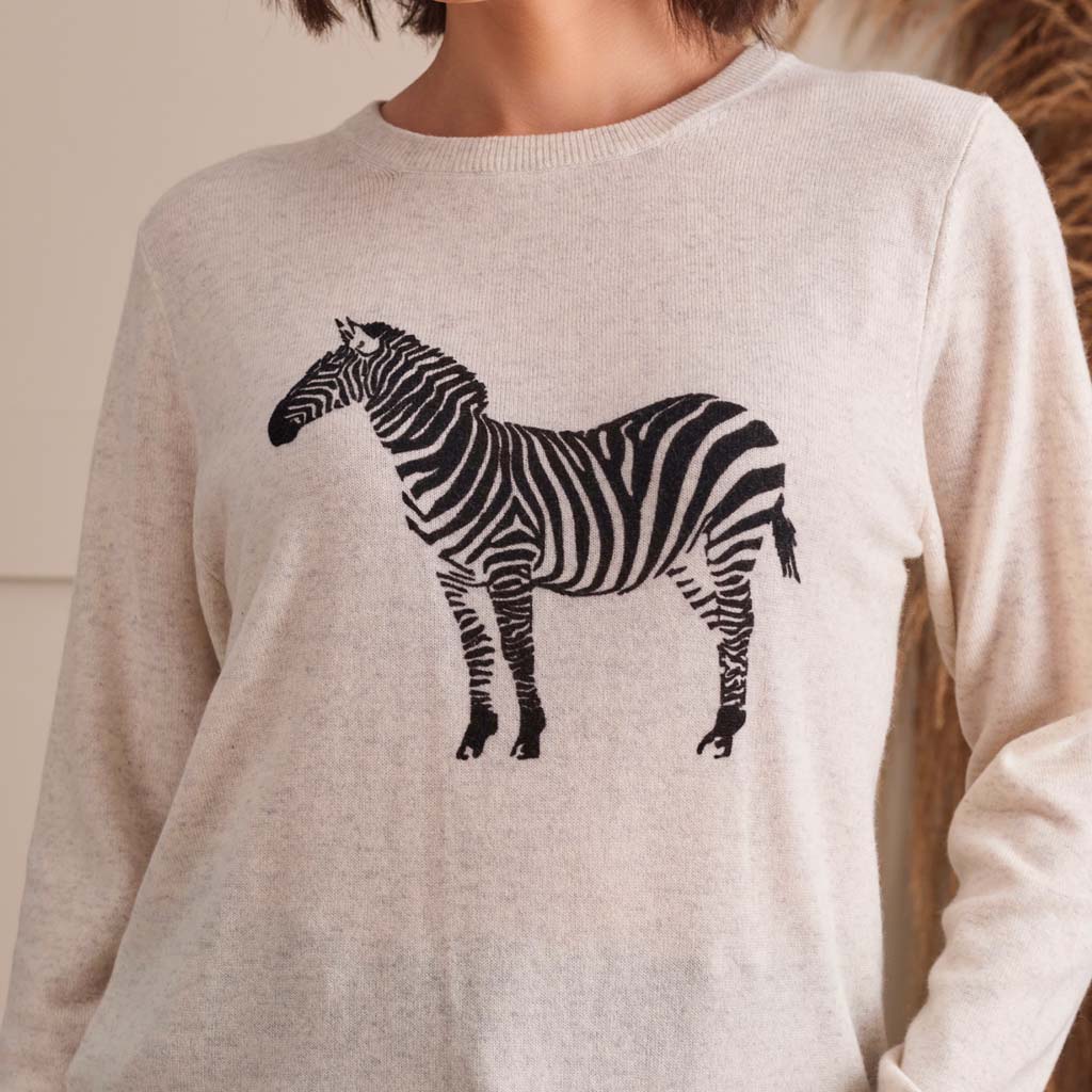 A person wearing a light beige crewneck sweater with a black and white zebra graphic printed on the front, set against a softly blurred neutral background.