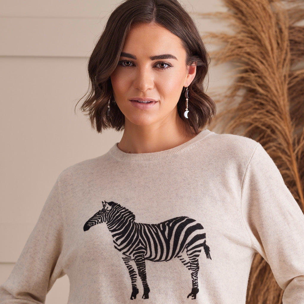 A woman wearing a light beige crewneck sweater with a black and white zebra graphic on the front, posed against a neutral indoor background with dried pampas grass.