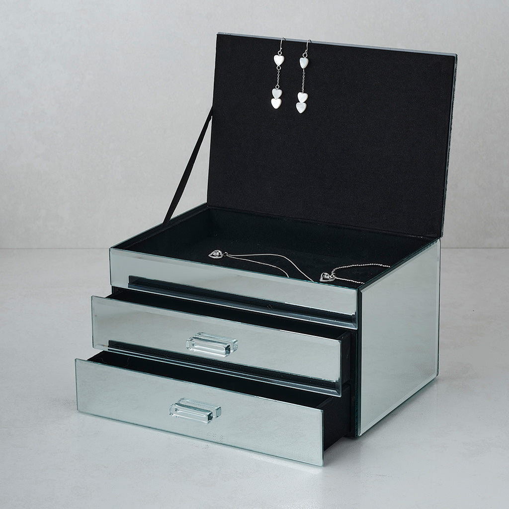 A rectangular mirrored jewelry box with three drawers, shown open with black velvet interior lining and jewelry displayed inside, including a pair of dangling earrings and necklaces.