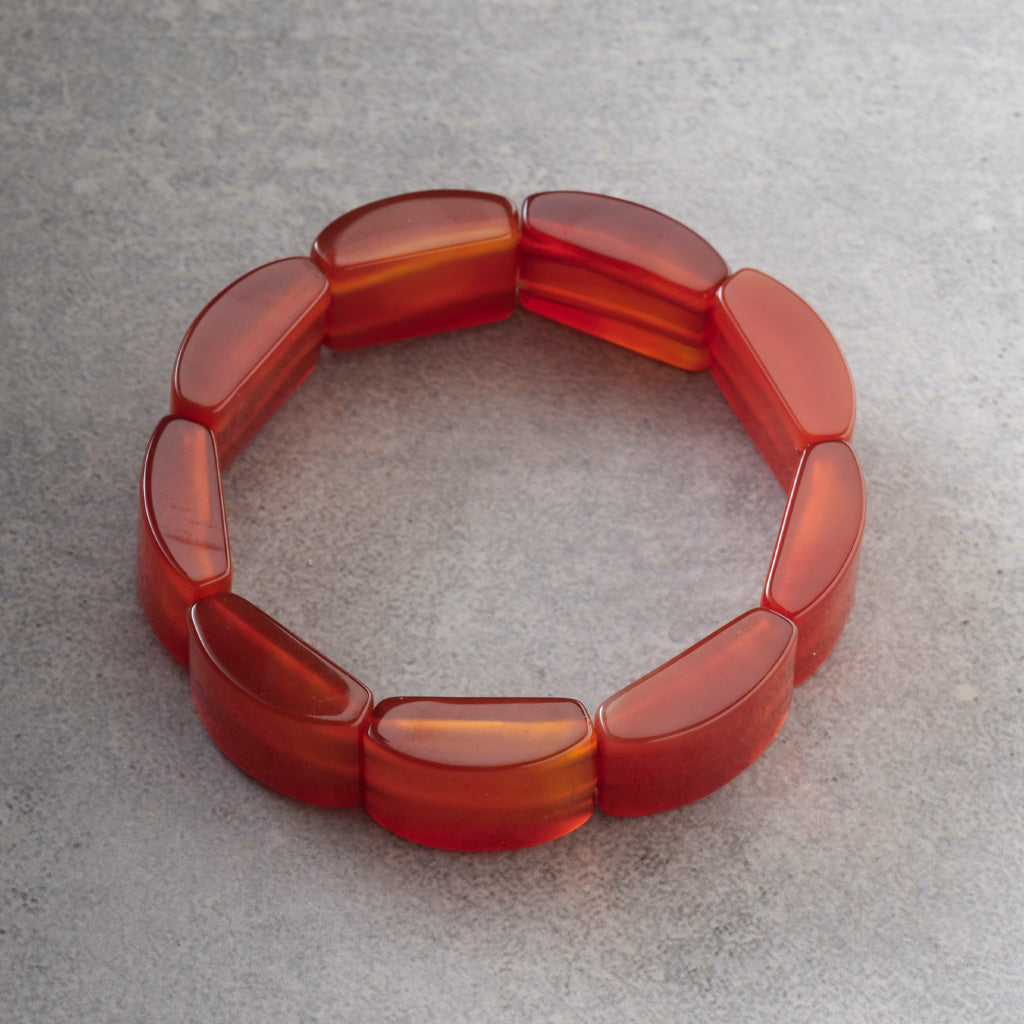 A translucent red-orange beaded stretch bracelet made of rounded, polished segments, displayed on a textured gray surface.
