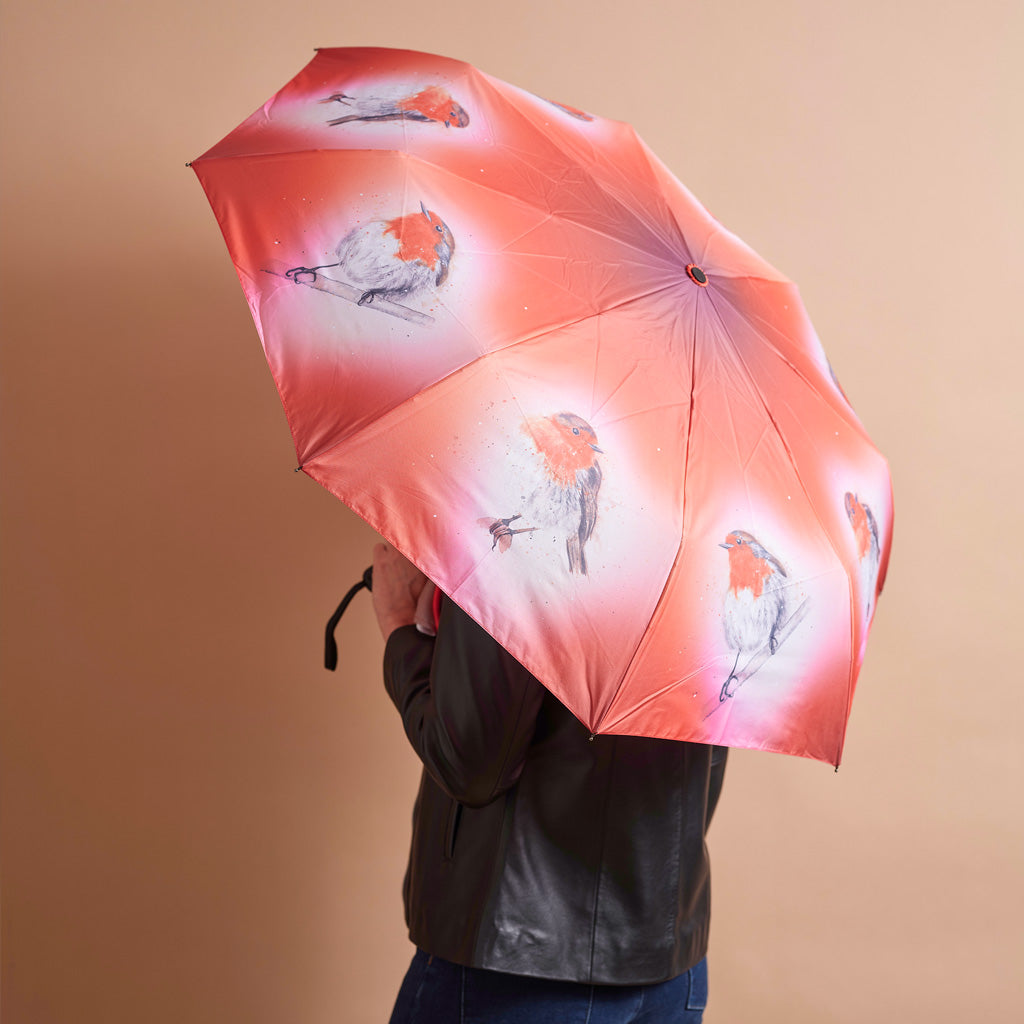 A person holding an open umbrella with a peach-pink background and bird illustrations. The person is wearing a black leather jacket and blue jeans, shown from the back and side against a solid beige background.