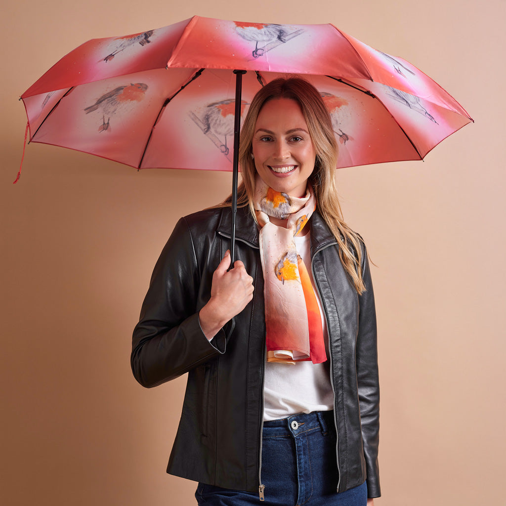 A woman holding a pink umbrella with bird illustrations, wearing a black leather jacket, white t-shirt, and blue jeans against a solid beige background.