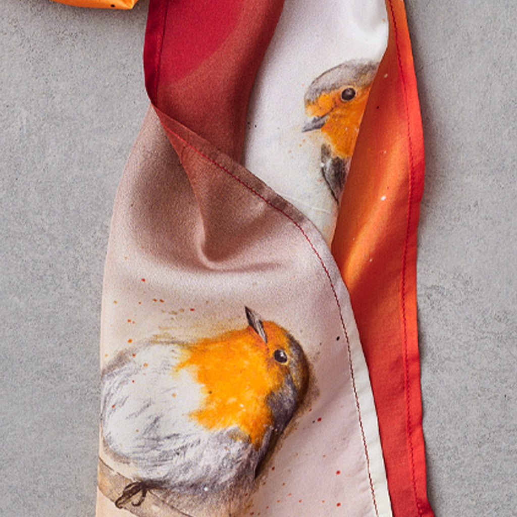 A close-up of a silk scarf featuring a watercolor-style robin bird print and a red-orange gradient border, laid on a gray textured surface.