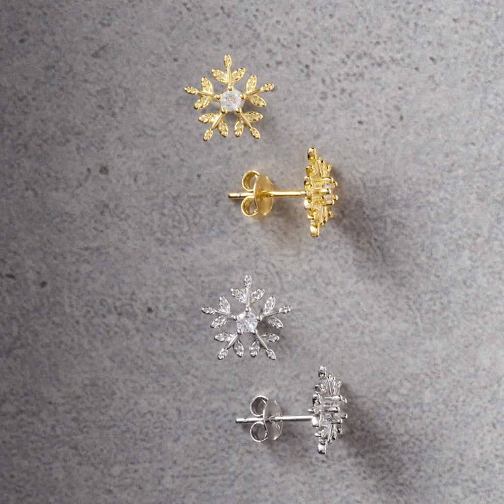 Four snowflake-shaped stud earrings arranged on a textured gray surface, including two gold-toned earrings with clear stones and two silver-toned earrings with clear stones.