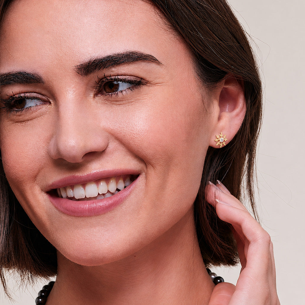 A pair of Gold-Plated snowflake stud earrings, with one shown from the front featuring a clear round-cut center stone, and the other showing the back with a post and butterfly clasp, set against a textured gray background.