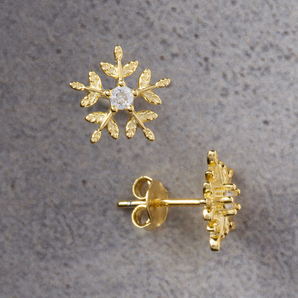 A pair of gold snowflake stud earrings, with one shown from the front featuring a clear round-cut center stone, and the other showing the back with a post and butterfly clasp, set against a textured gray background.