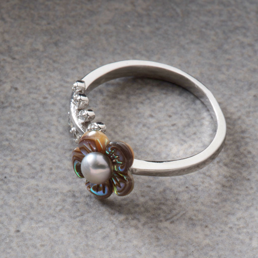 A silver ring featuring a flower-shaped setting with iridescent brown and blue-green mother-of-pearl petals, a central white pearl, and small clear stones along the band, shown on a textured gray surface.